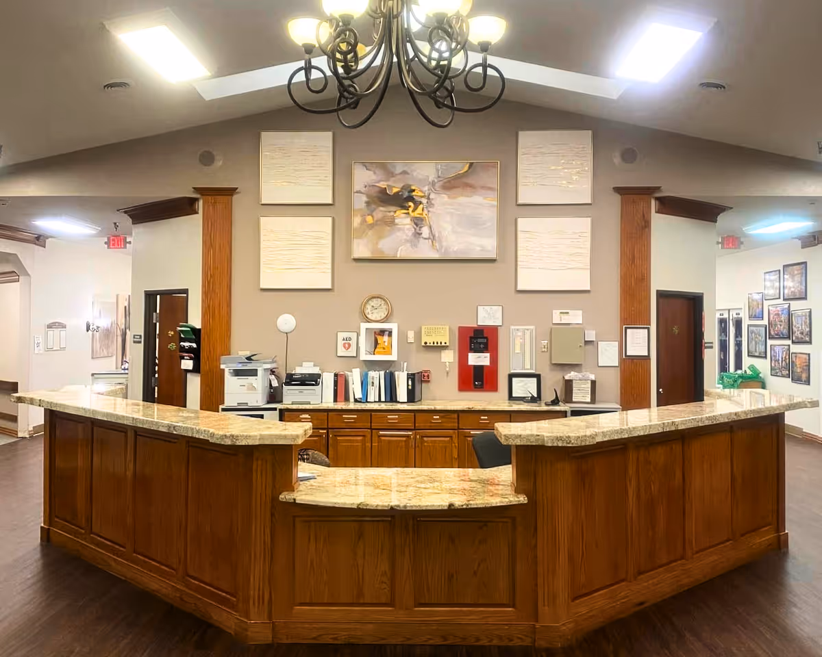 Reception area inside Whispering Pines Lodge featuring a large wooden front desk with a marble countertop. Behind the desk are office supplies, a printer, binders, a clock, and a red fire alarm panel mounted on the beige wall. Above the desk are four framed artworks, including an abstract painting in the center. The ceiling has two skylights and a decorative chandelier. Hallways with doors and framed pictures are visible on either side of the reception area.