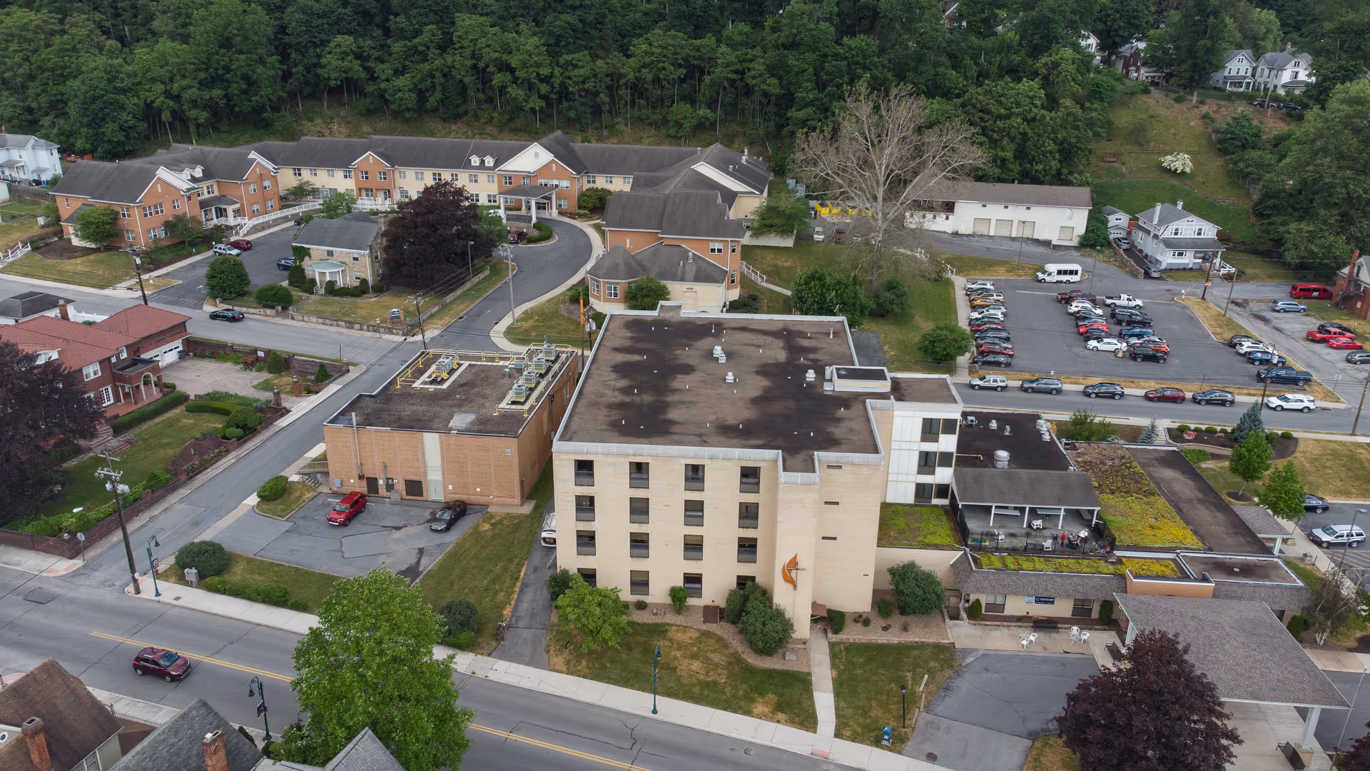 Aerial view of Cedarwood Rehabilitation and Healthcare Center showing multiple connected buildings with flat roofs, surrounding parking lots, and nearby residential houses with trees and greenery.