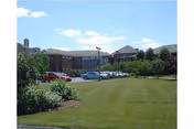 Exterior view of a large brick building with multiple windows, surrounded by a well-maintained lawn and several parked cars under a blue sky with a few clouds.