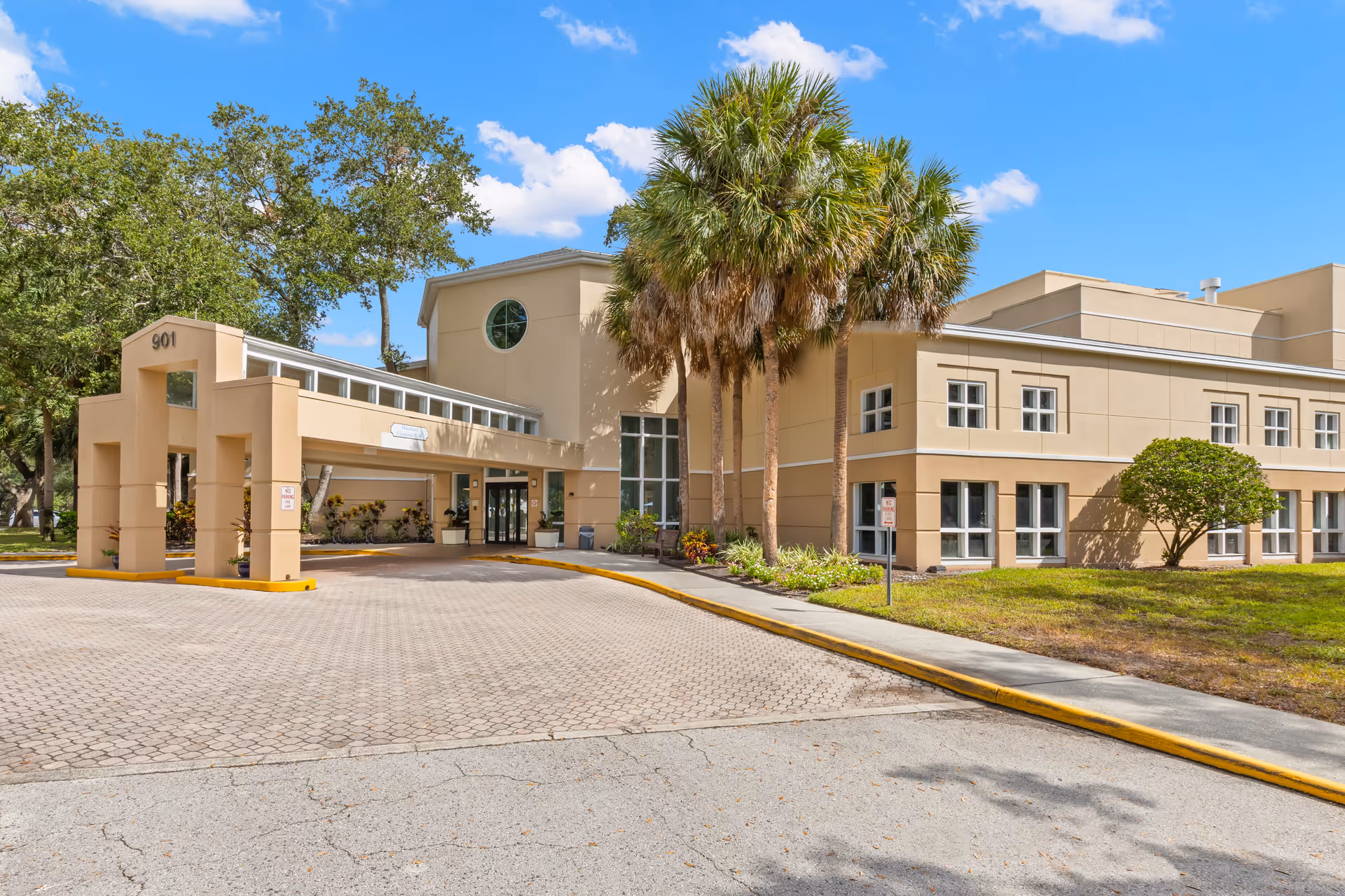 Exterior view of a beige senior living facility building with a covered entrance, palm trees, and a paved driveway under a blue sky with some clouds.