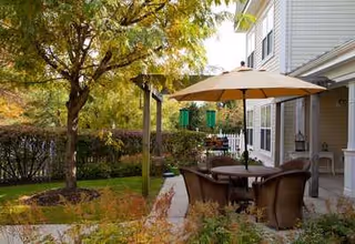 Outdoor patio area with a table, umbrella and chairs beside a building and landscaped yard with trees.