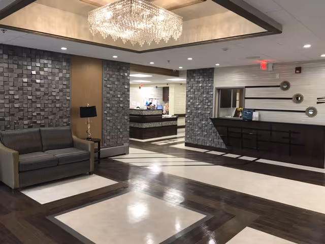 Interior view of a senior living facility lobby with a modern chandelier, a gray couch with a side table and lamp, textured wall panels, and a reception desk in the background.