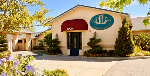 Front entrance of a single-story senior living building with a red awning over glass double doors, an oval sign on the facade, and landscaped shrubs and flowers.