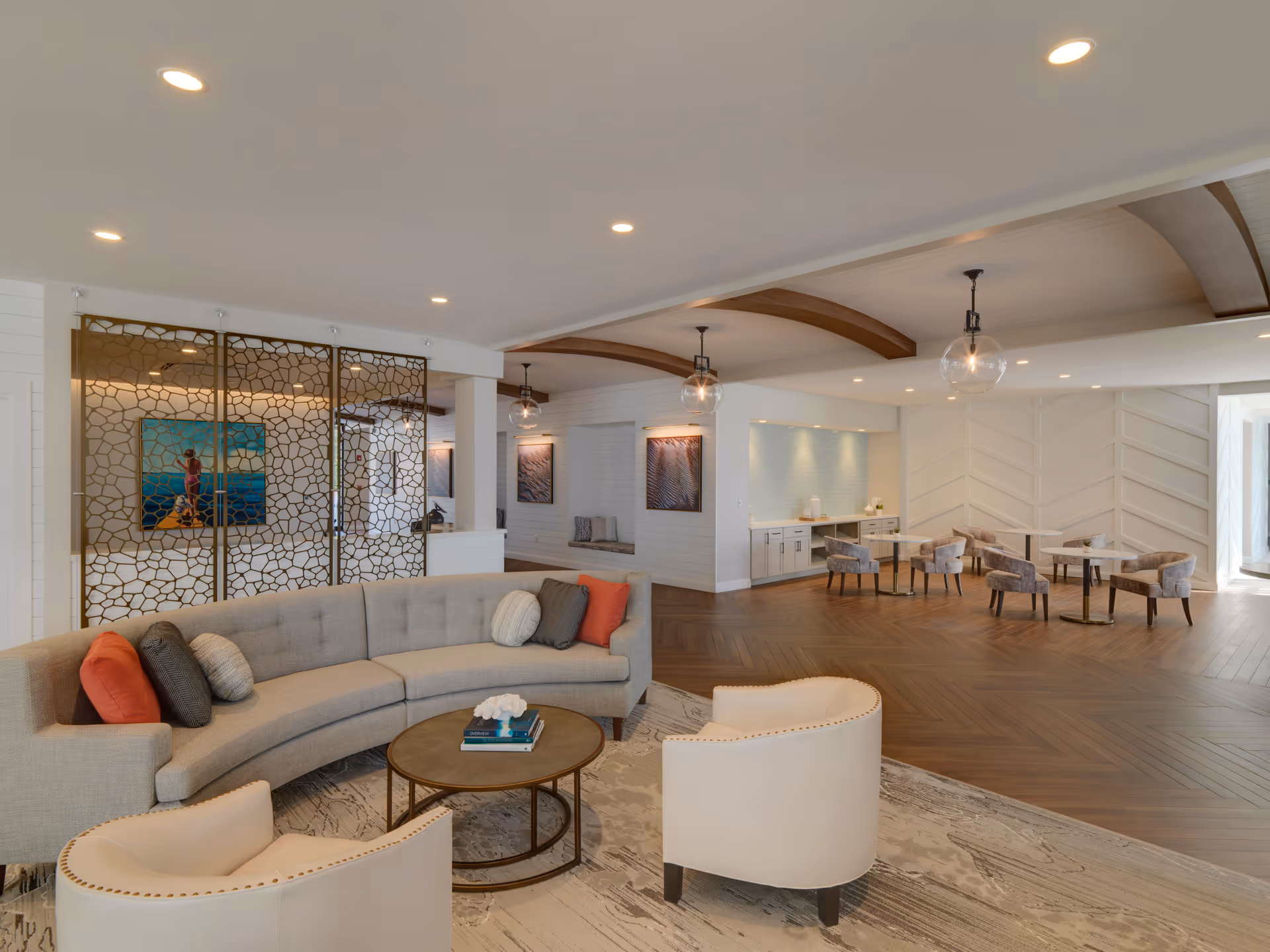 A spacious and modern living area featuring a curved beige sofa with multiple cushions, two cream-colored armchairs, and a round coffee table with books and a decorative item. The room has wooden flooring with a patterned rug, decorative ceiling beams, and pendant lights with glass globes. In the background, there are small tables with upholstered chairs and a built-in counter with cabinets and a light blue tiled backsplash. Decorative wall panels and artwork are also visible.