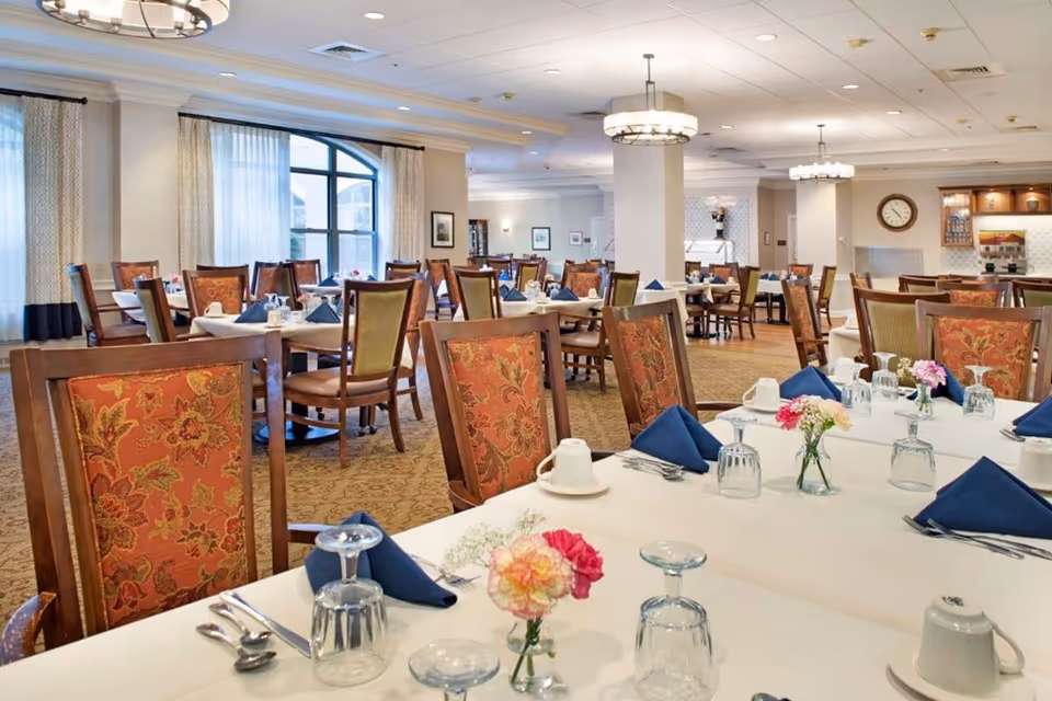 A spacious and well-lit dining room with multiple tables set for a meal. Tables are covered with white tablecloths, adorned with small floral centerpieces, blue folded napkins, upside-down glasses, cups, and silverware. The room features patterned upholstered chairs, large windows with curtains, and ceiling lights. A clock and framed pictures are visible on the walls in the background.