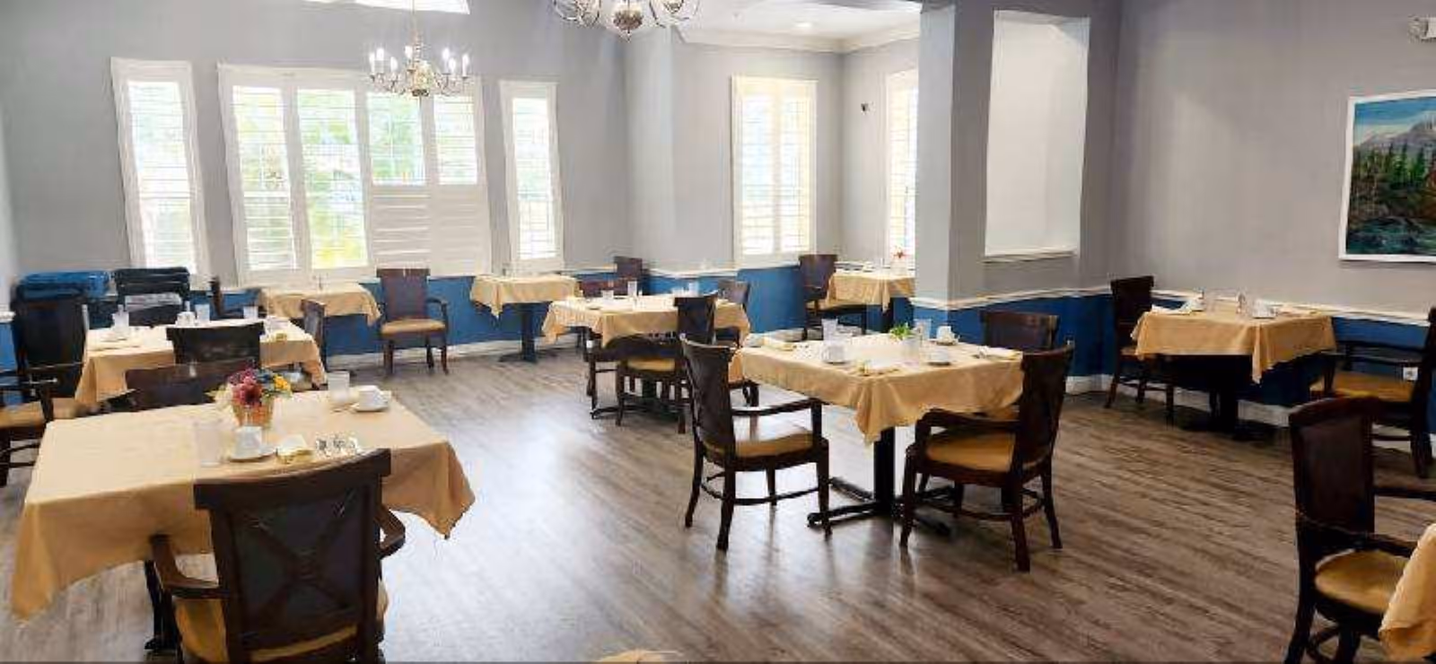A bright dining room with several tables covered in beige tablecloths, each set with cups, glasses, and small flower arrangements. The room has large windows with white shutters, wooden chairs with cushioned seats, light gray walls with a blue lower trim, and a wooden floor. A chandelier hangs from the ceiling, and a landscape painting is visible on one wall.