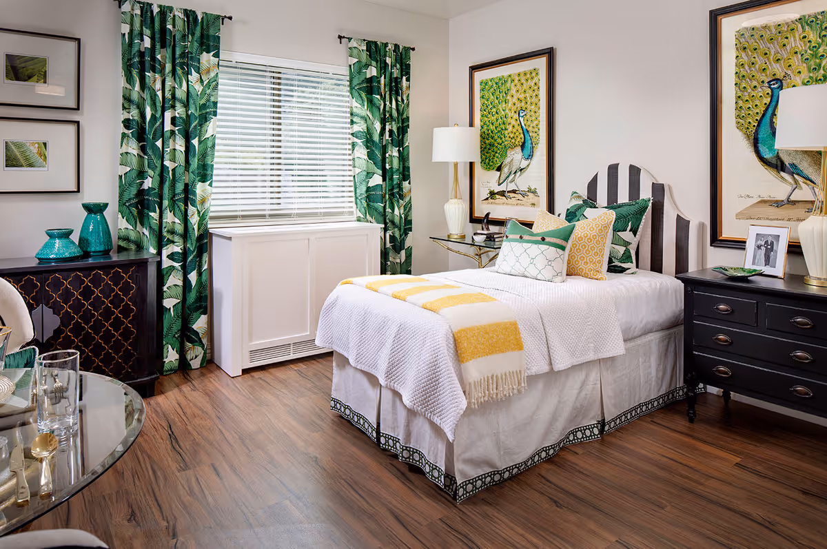 A cozy bedroom with a single bed dressed in white bedding and yellow and green accent pillows and a yellow striped throw blanket. The room features wooden flooring, green leafy patterned curtains, two framed peacock artworks on the walls, a black dresser with a lamp and photo frame, and a small black cabinet with decorative green vases.