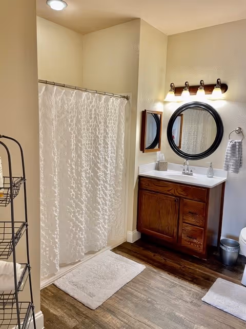 A bathroom with a white textured shower curtain, a wooden vanity with a white countertop, a round mirror above the sink, a wall-mounted light fixture with four bulbs, a towel ring holding a gray towel, a metal shelving unit with towels, and a white bath mat on a wood-look floor.