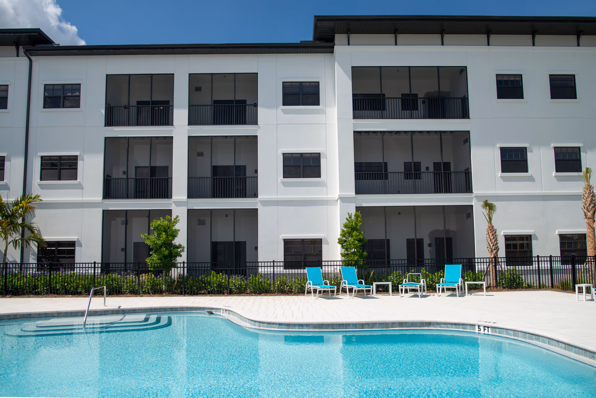 Outdoor swimming pool with clear blue water in front of a modern three-story white building with balconies and windows. Several turquoise lounge chairs and small tables are arranged on the pool deck. Palm trees and green shrubs line the area between the pool and the building.