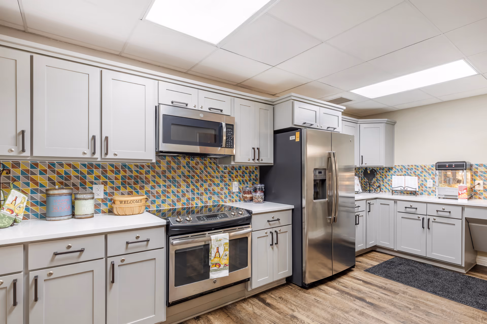A modern kitchen with white cabinets, a stainless steel microwave and oven, a stainless steel refrigerator, and a colorful geometric tile backsplash. The kitchen has wooden flooring and bright overhead lighting. Various kitchen items such as containers, a welcome basket, and a popcorn machine are placed on the countertops.