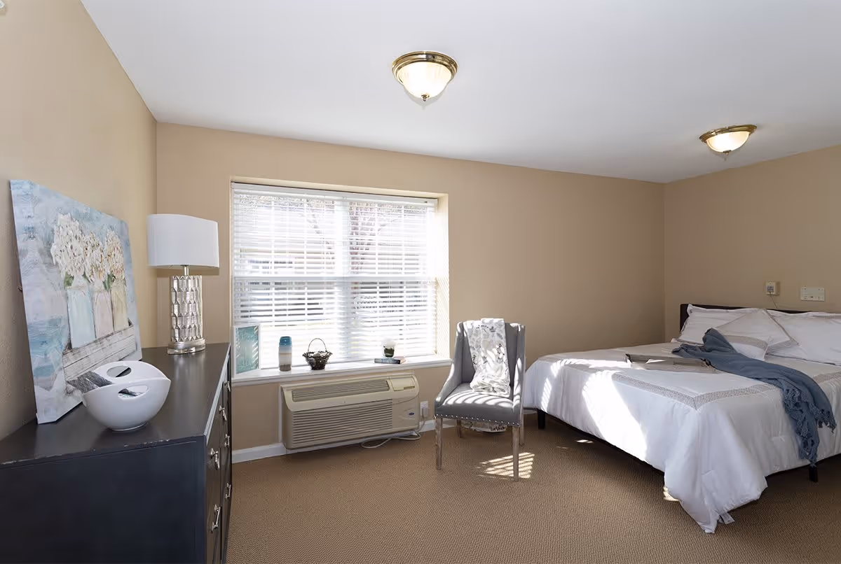 A bright bedroom with beige walls and carpet. The room features a large window with white blinds, a bed with white bedding and a blue throw, a gray upholstered chair with a patterned blanket, and a dark dresser with a lamp, decorative bowls, and a floral painting resting on it. An air conditioning unit is installed below the window.