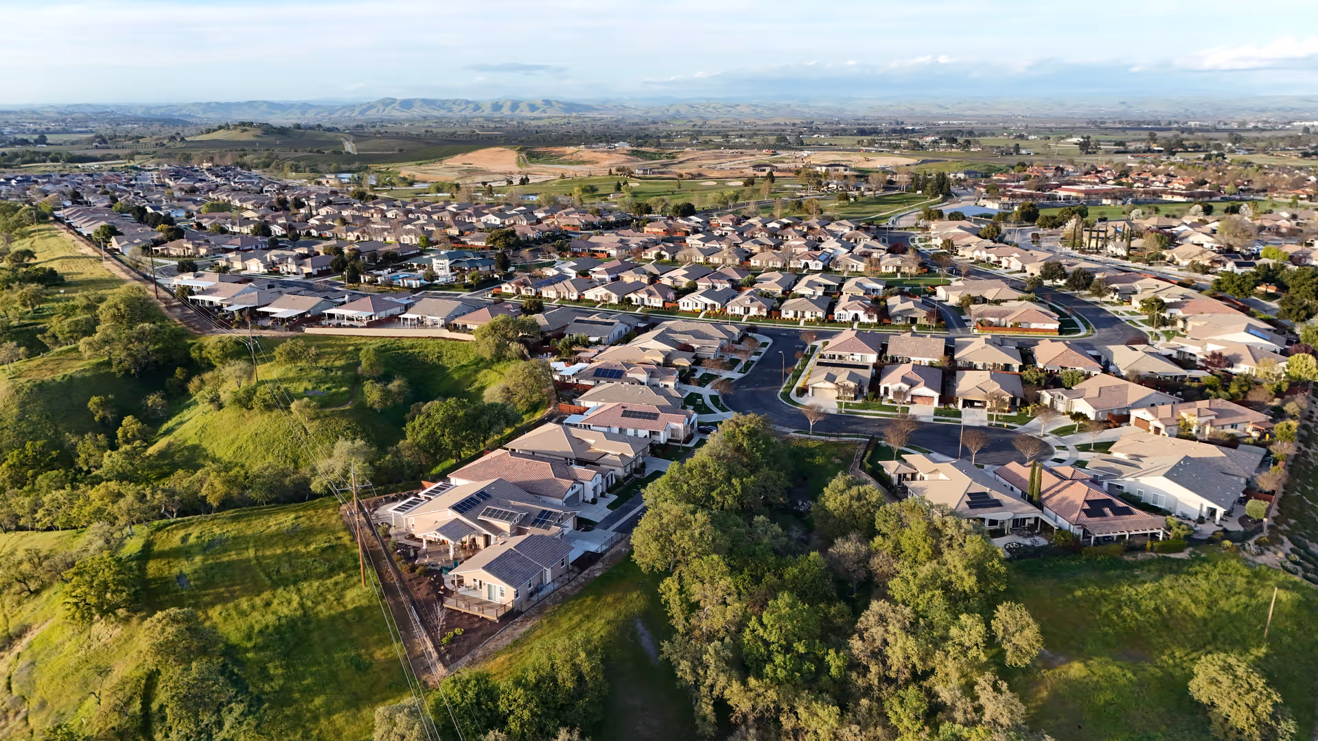 Aerial view of a single-story residential community clustered along curving roads next to green hills and trees.