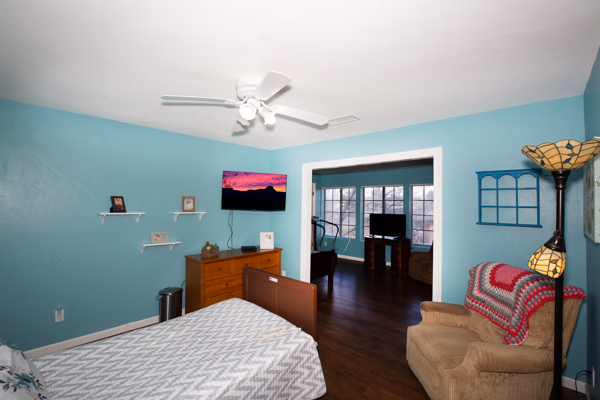 A cozy bedroom with teal walls, a bed with a gray and white chevron patterned blanket, a wooden dresser with a flat screen TV mounted above it, and small shelves with framed photos. There is an armchair with a red and gray crocheted blanket draped over it, next to a floor lamp with a stained glass shade. The room opens into a living area with large windows and another TV visible.