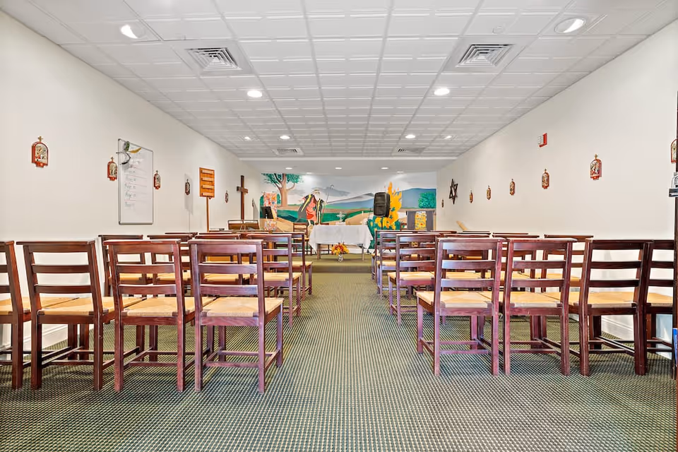 Interior view of a small chapel or worship room with rows of wooden chairs facing an altar at the front. The altar is decorated with a white cloth, flowers, and religious symbols including a cross on the wall and a mural depicting a biblical scene. The room has a carpeted floor and white walls with religious plaques mounted on them.