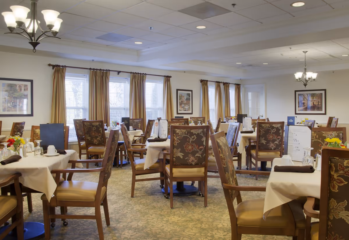 A well-lit dining room with multiple tables covered in white tablecloths, each set with menus, glasses, cups, and napkins. The chairs have floral patterned upholstery and wooden frames. Large windows with striped curtains allow natural light to fill the room. The walls are decorated with framed artwork, and there are two ceiling light fixtures providing additional lighting.