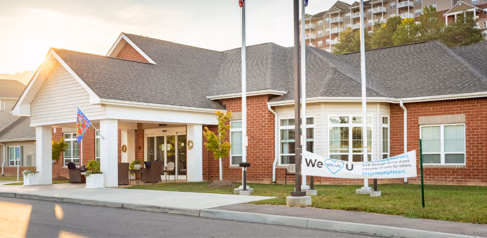 Exterior view of a single-story brick building with a covered entrance and seating area. There is a colorful flag on a pole near the entrance and a banner on the lawn that reads 'We VALUE U' with hashtags #UnityHeroes and #HarmonyHeart. The sun is setting in the background, casting a warm light over the scene.