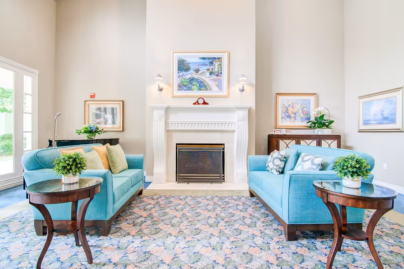 A bright and elegant living room with two turquoise sofas facing each other across a floral patterned rug. Each sofa has decorative pillows, and there are small round wooden side tables with potted plants on either side. A white fireplace with a mantel is centered on the far wall, above which hangs a colorful painting. Additional framed artwork and plants decorate the walls and furniture around the room. Large windows on the left side allow natural light to fill the space.
