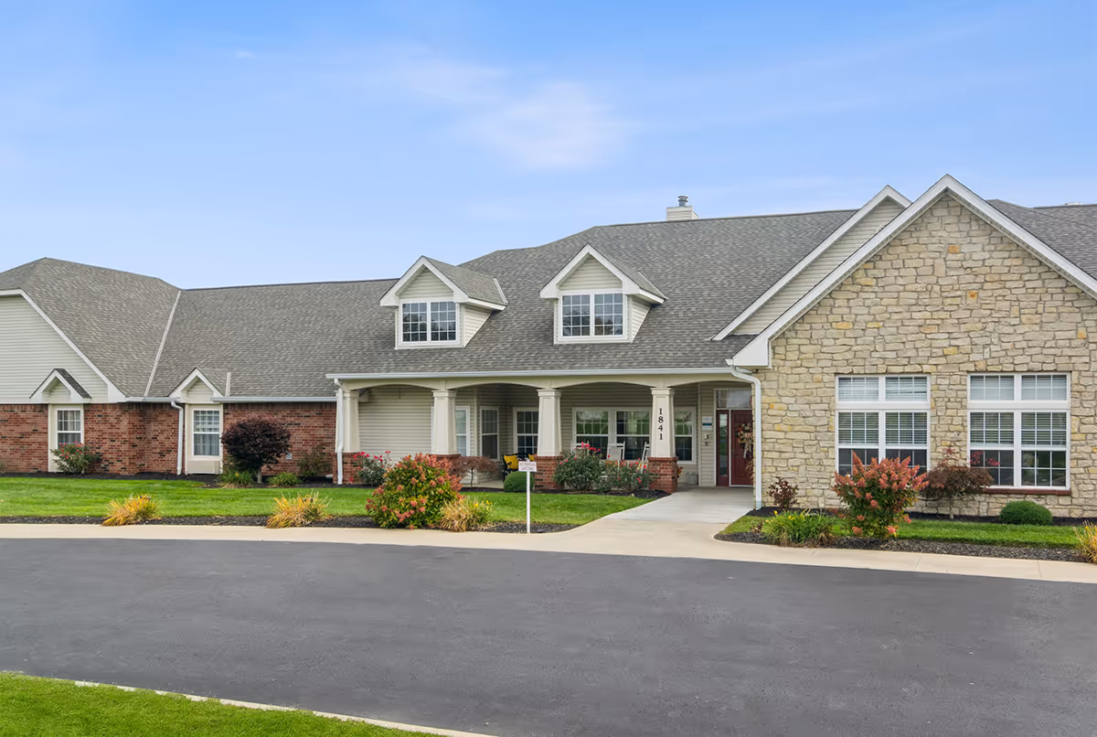 Exterior front view of a single-story senior living facility building with a combination of brick, stone, and siding. The building features a covered entrance with columns, multiple windows, and a well-maintained lawn with shrubs and flowers under a clear blue sky.