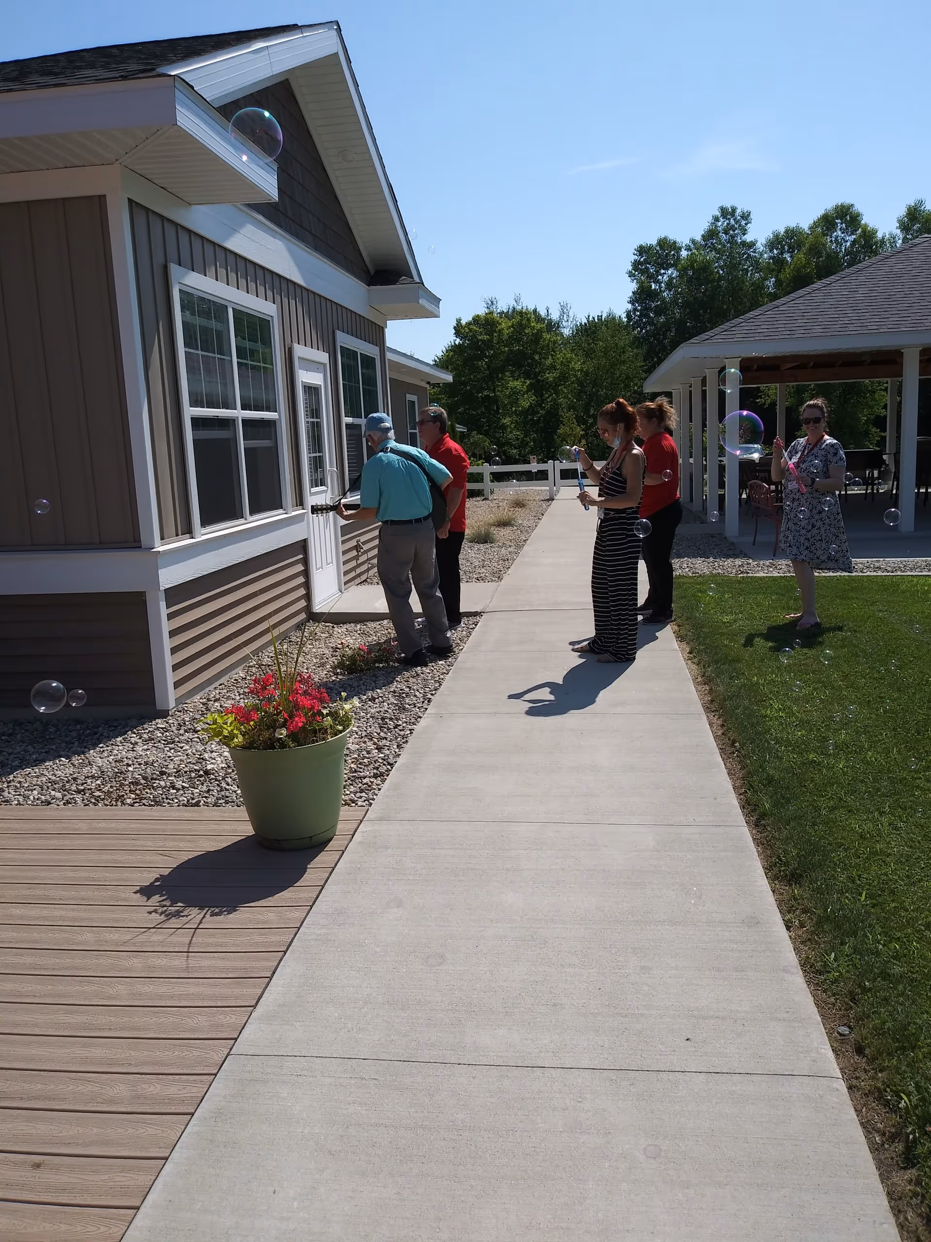 Residents and staff stand on a sidewalk outside a single-story assisted living building with a potted plant and a covered patio under a clear sky.