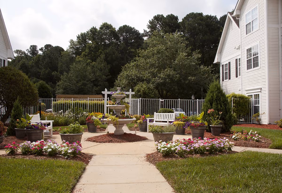 Outdoor garden area at Brighton Gardens of Raleigh featuring a central multi-tiered fountain surrounded by flower beds and potted plants. White benches are placed around the fountain, with a white fence and trees in the background. The side of a white building is visible on the right.