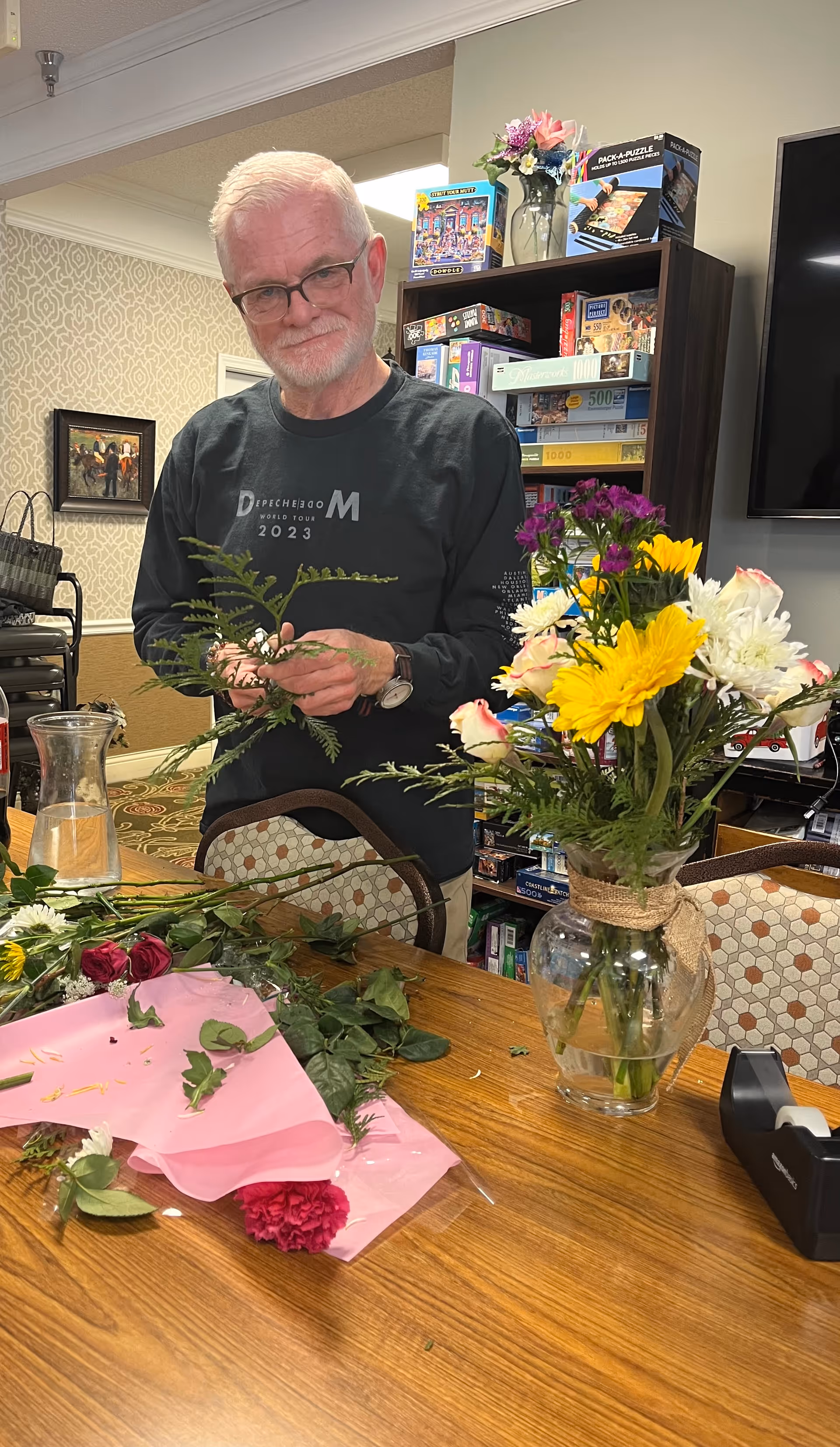 An elderly man with white hair and glasses arranging flowers at a wooden table inside a room. The table has various flowers, leaves, and a pink wrapping paper. Behind him is a bookshelf filled with board games and puzzles, and a large TV mounted on the wall to the right.