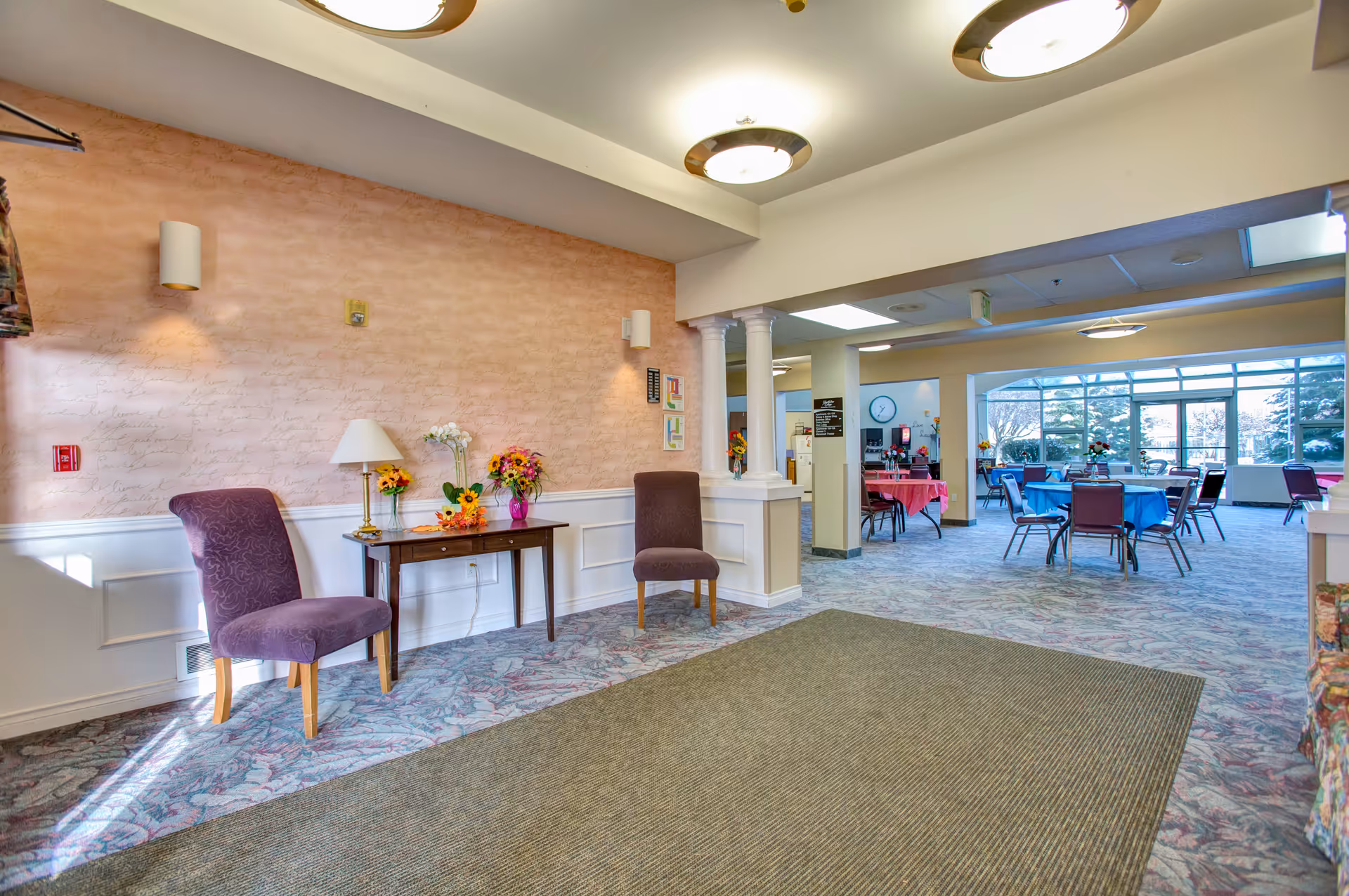 Interior view of a senior living facility common area with two purple chairs and a small table with flowers and a lamp against a peach-colored wall. The area opens into a larger dining room with tables covered in blue and pink tablecloths and chairs, with large windows letting in natural light.