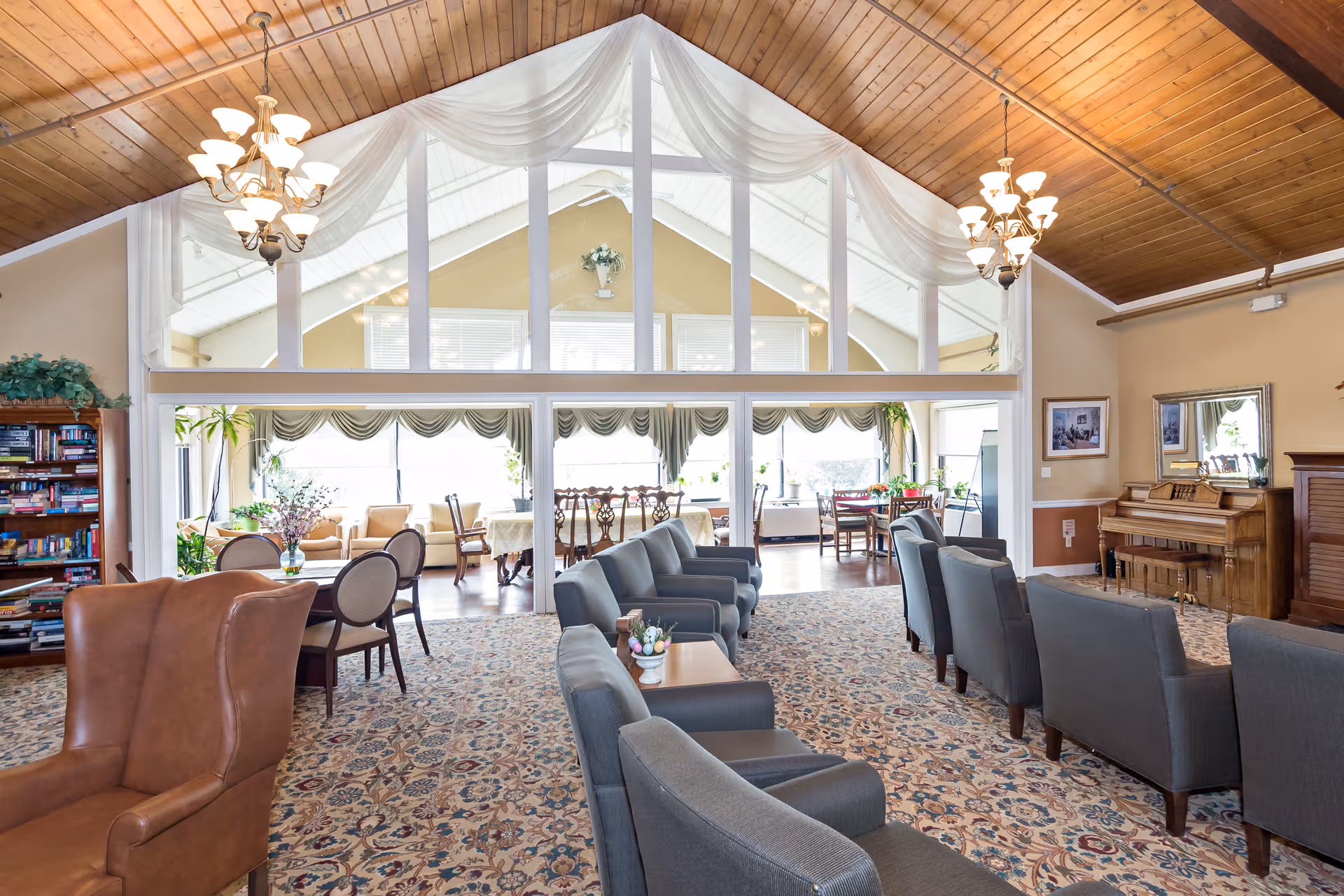 Spacious, well-lit assisted living common room with rows of armchairs, a piano, chandeliers and a vaulted wood ceiling overlooking a bright sunroom.