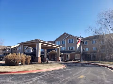 Exterior view of a multi-story senior living facility building with a covered entrance driveway, a flagpole with a flag, and some bushes and trees around the property under a clear blue sky.
