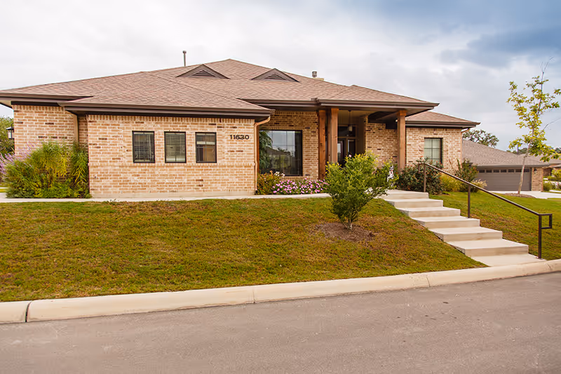 Front exterior view of a single-story brick building with a sloped roof, a small set of stairs with a handrail leading to the entrance, and landscaping including grass, bushes, and a small tree.