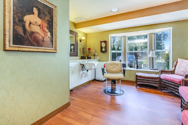 A bright interior room with a salon chair and sink, wicker seating by a bay window, and framed artwork on the wall.