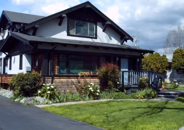 Exterior view of a two-story residential-style building with white walls and dark wooden trim, surrounded by a well-maintained garden with green grass, bushes, and flowers under a partly cloudy sky.