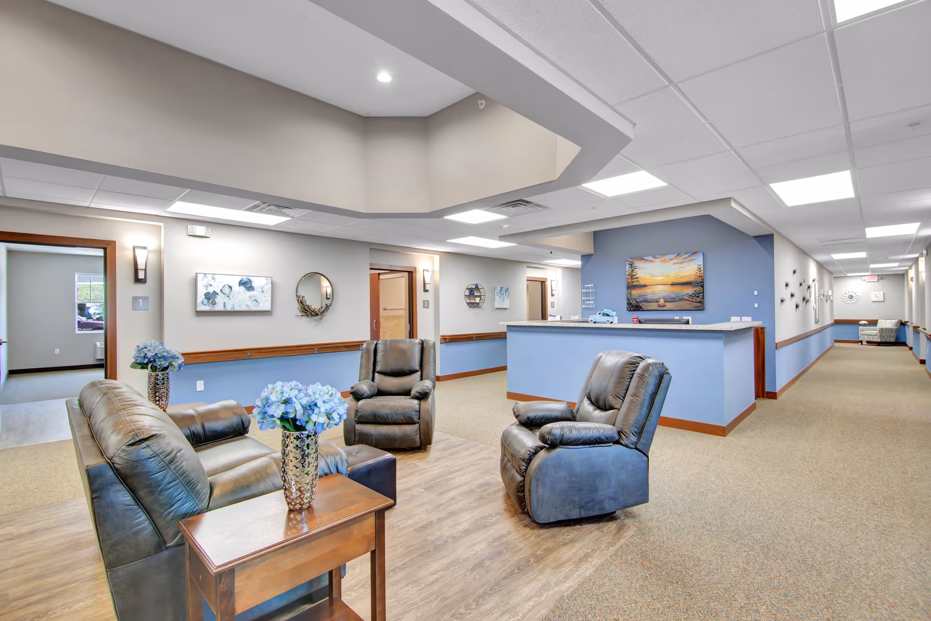 A bright and spacious senior living facility common area with comfortable leather seating including a sofa and two armchairs arranged around a wooden side table with a vase of blue flowers. The walls are painted in soft beige and blue tones, decorated with artwork and a round mirror. A reception desk is visible in the background along with a long hallway leading to other rooms.