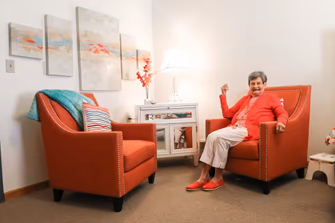 An elderly woman wearing a red cardigan and white pants sits comfortably in an orange armchair in a cozy living room. Another matching orange armchair with a striped pillow and a teal blanket is nearby. A white side table with a lamp and decorative items is between the chairs, and abstract artwork hangs on the wall behind them.