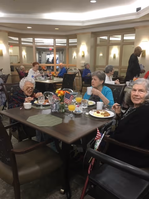 Several elderly residents dine at a communal dining room table in a senior living facility, with plates, drinks, small American flags, and flowers on the table.