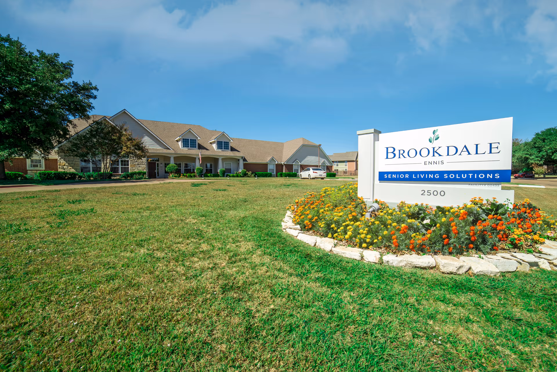 Exterior view of Brookdale Ennis senior living facility with a large lawn, flower bed, and a white sign displaying the facility name and address under a blue sky.