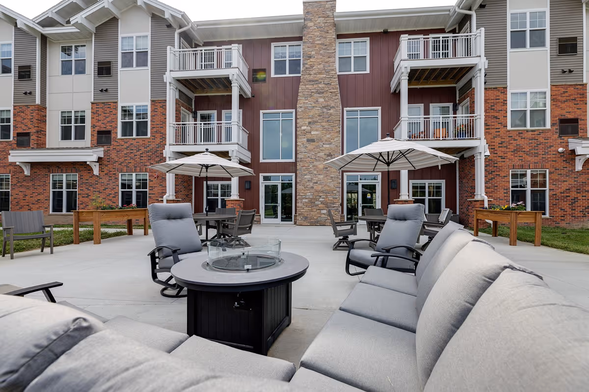 Outdoor patio with cushioned seating, umbrellas, and tables in front of a multi-story brick and siding senior living building.