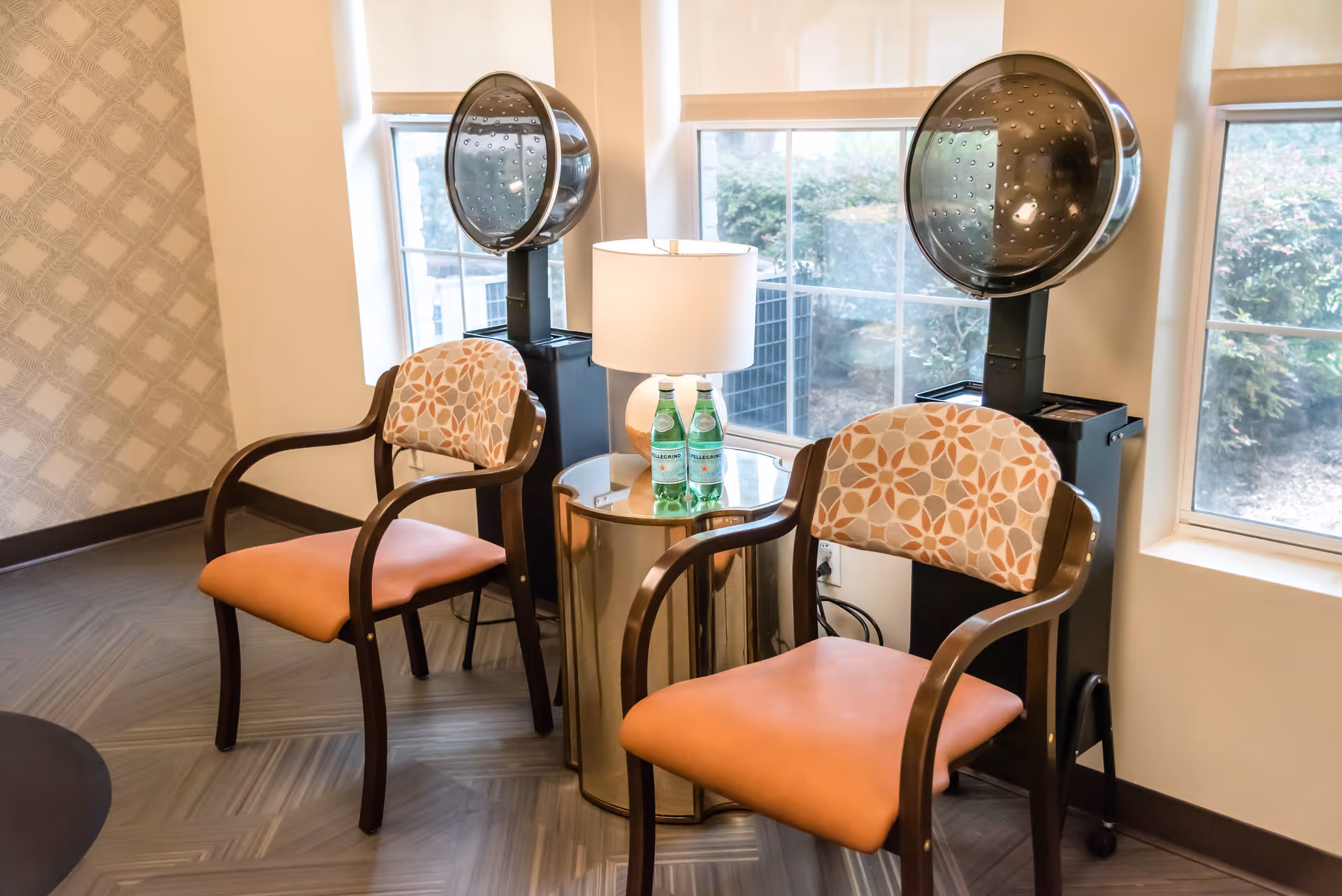 Two patterned chairs with orange seats are placed on either side of a small round table with a lamp and two bottles of sparkling water. Behind each chair is a vintage hair dryer. The setting is next to large windows with a view of greenery outside.