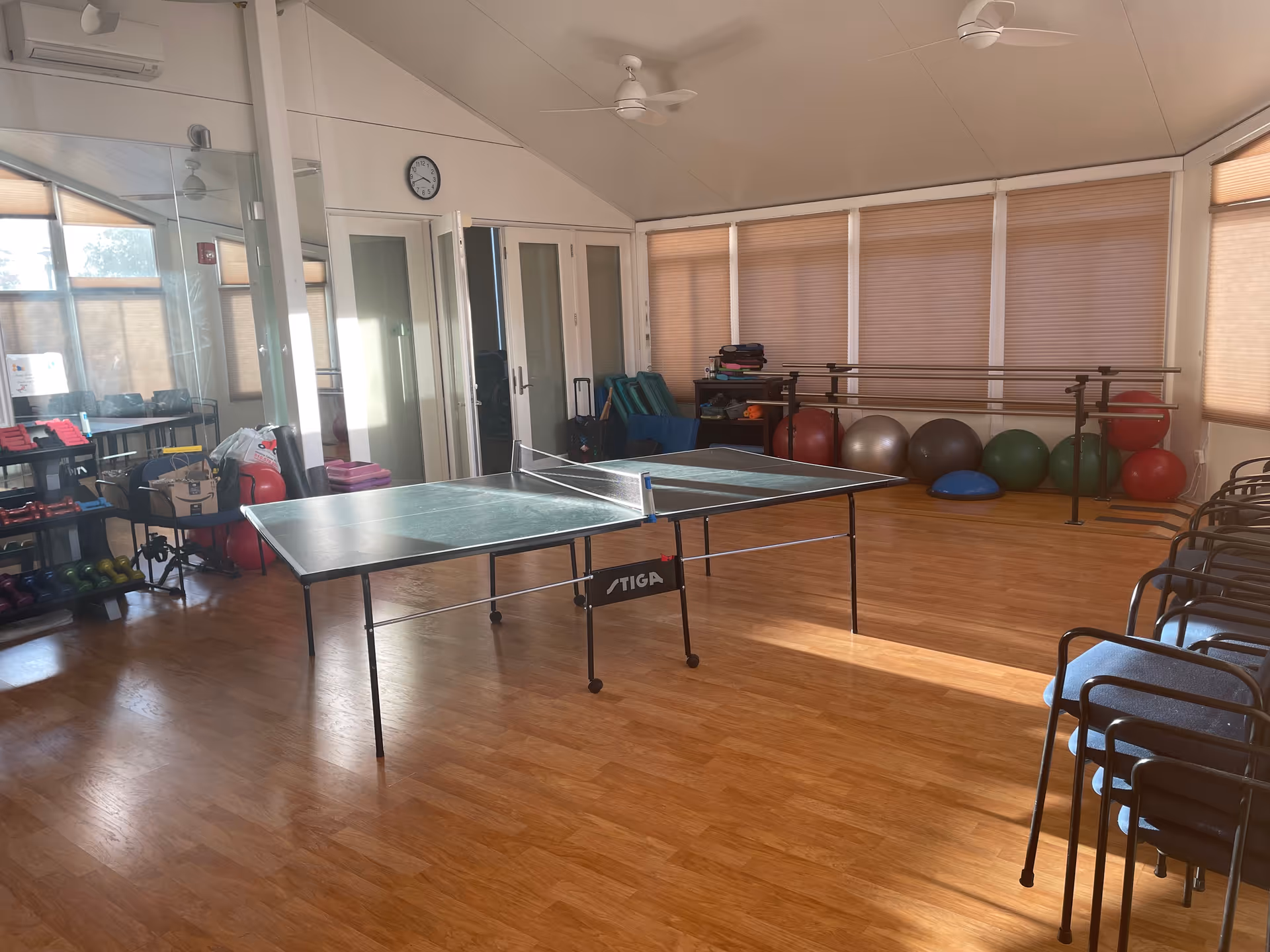 A well-lit activity room with a ping pong table in the center, exercise balls lined up against the far wall, stacked chairs on the right, and shelves with various exercise equipment on the left. The room has wooden flooring, large windows with blinds, ceiling fans, and a clock on the wall above double doors.