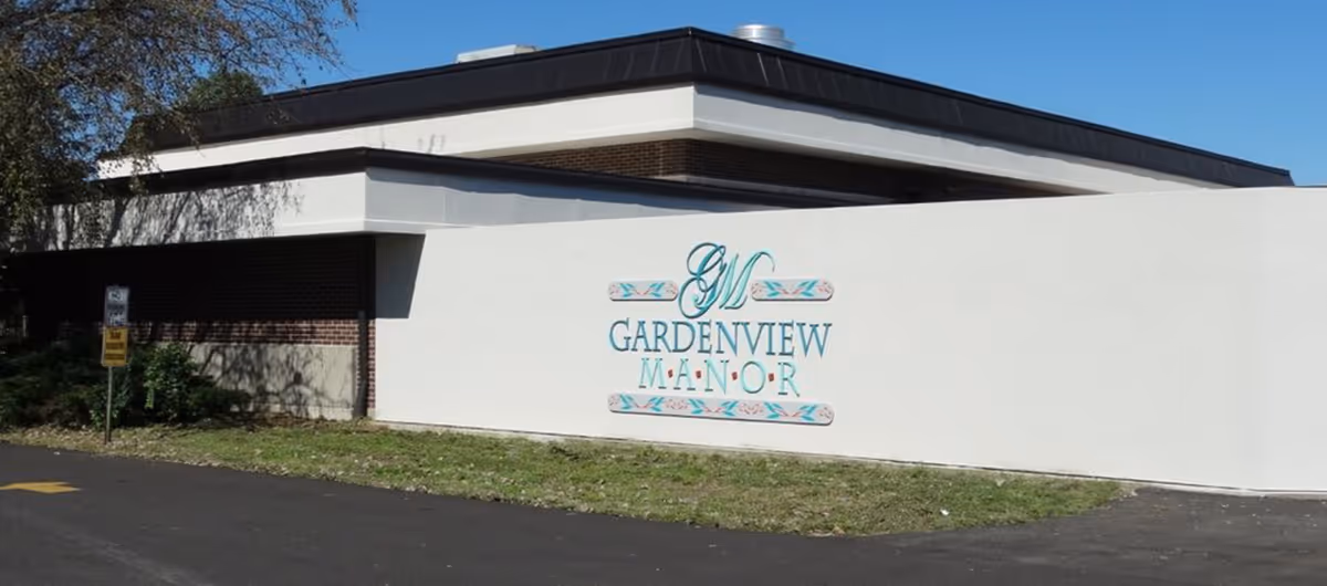 Exterior view of a building with a white wall displaying the sign 'Gardenview Manor' in decorative lettering. The building has a flat roof and brick accents, with a paved driveway or parking area in front and some greenery on the side.