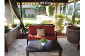 Outdoor covered patio area with a red cushioned loveseat, a small black coffee table with a potted plant, and large potted plants on the side. The patio overlooks a landscaped garden with trimmed bushes and trees.