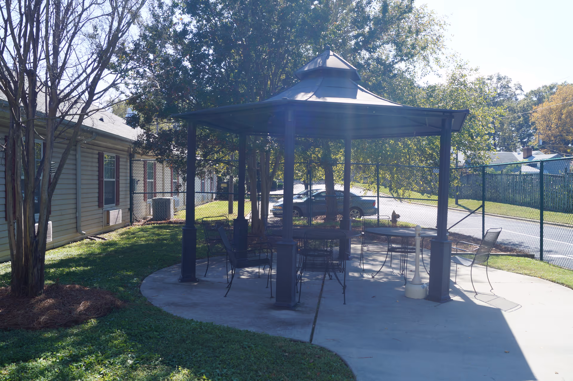A metal gazebo with tables and chairs on a concrete patio beside a residential building and fenced parking area.