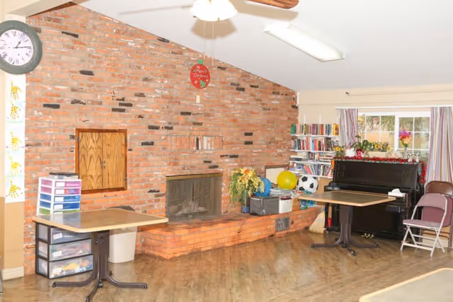A cozy room with a brick fireplace on the left wall, two tables with chairs, a black piano decorated with flowers, a bookshelf filled with books, and a window with striped curtains letting in natural light.