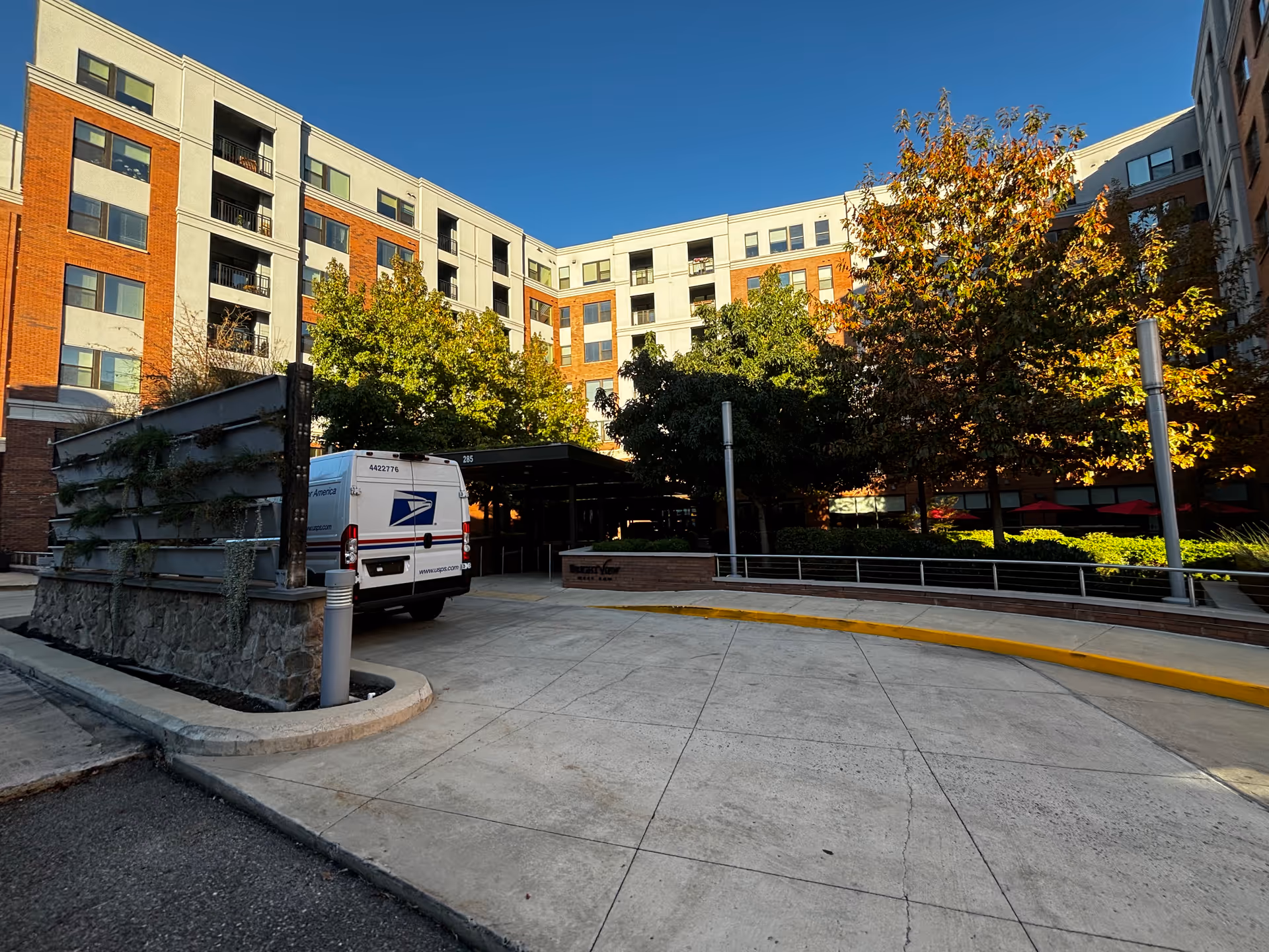 Exterior view of a multi-story senior living facility building with a driveway and a USPS delivery van parked near the entrance. The building has brick and light-colored paneling with multiple windows and balconies. Trees with autumn foliage are visible near the entrance under a clear blue sky.