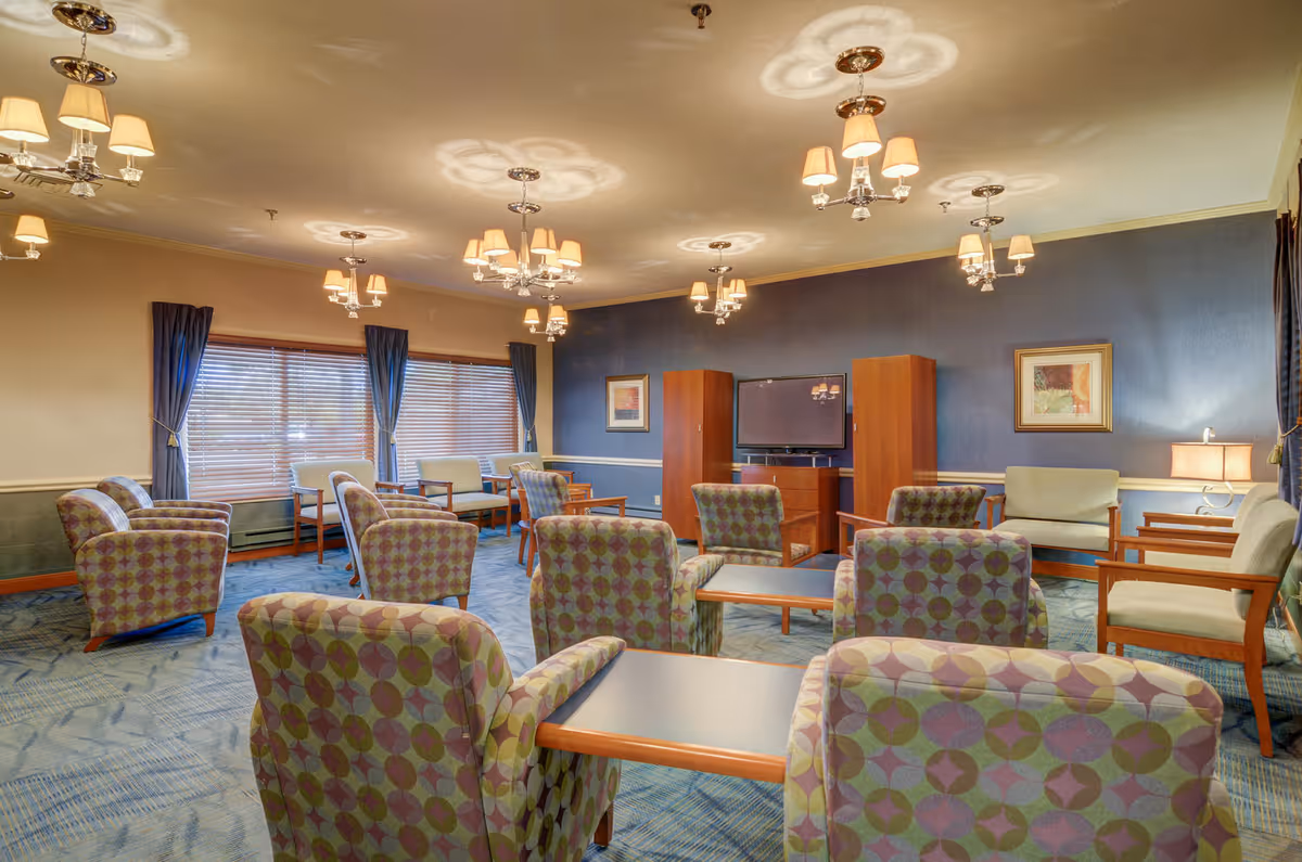 A spacious living room area with multiple patterned armchairs and wooden-framed chairs arranged around several tables. The room features blue walls, large windows with blue curtains, a flat-screen TV mounted between two wooden cabinets, framed artwork on the walls, and multiple ceiling light fixtures with lampshades.
