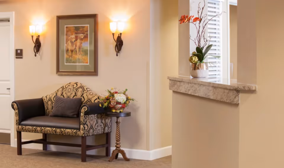 A cozy seating area in a senior living facility hallway featuring a patterned loveseat with a dark cushion, a small round wooden side table with a floral arrangement, a framed picture of two dogs on the wall, two wall-mounted light fixtures, and a window with white blinds and a decorative plant on the ledge.