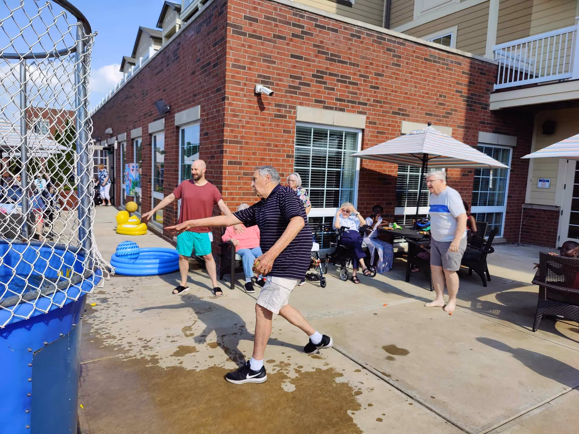 A group of elderly people and a younger man are enjoying an outdoor activity at a senior living facility. One elderly man is throwing a ball towards a dunk tank, while others watch and sit under umbrellas on a patio next to a brick building. There are inflatable pool toys and a small blue kiddie pool nearby.