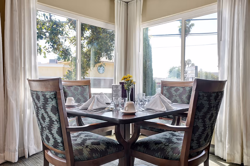 Sunlit dining table set for four with folded napkins, glassware, and a small flower centerpiece by large windows and curtains.