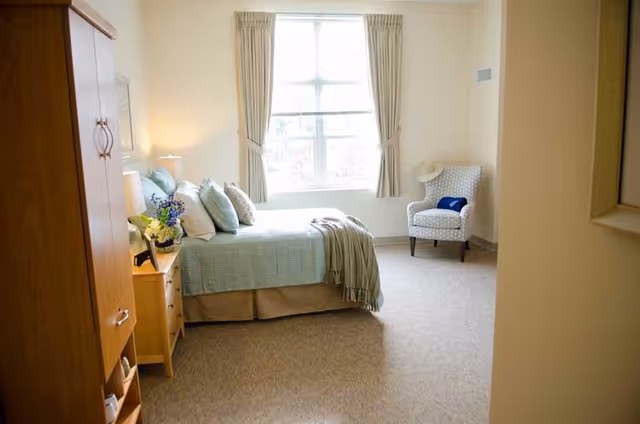 A bright and cozy bedroom with a large window covered by beige curtains. The room features a bed with light blue bedding and multiple pillows, a wooden nightstand with a lamp and flowers, a wooden wardrobe, and a patterned armchair with a hat and a blue item placed on it. The carpet is beige and the walls are light-colored.