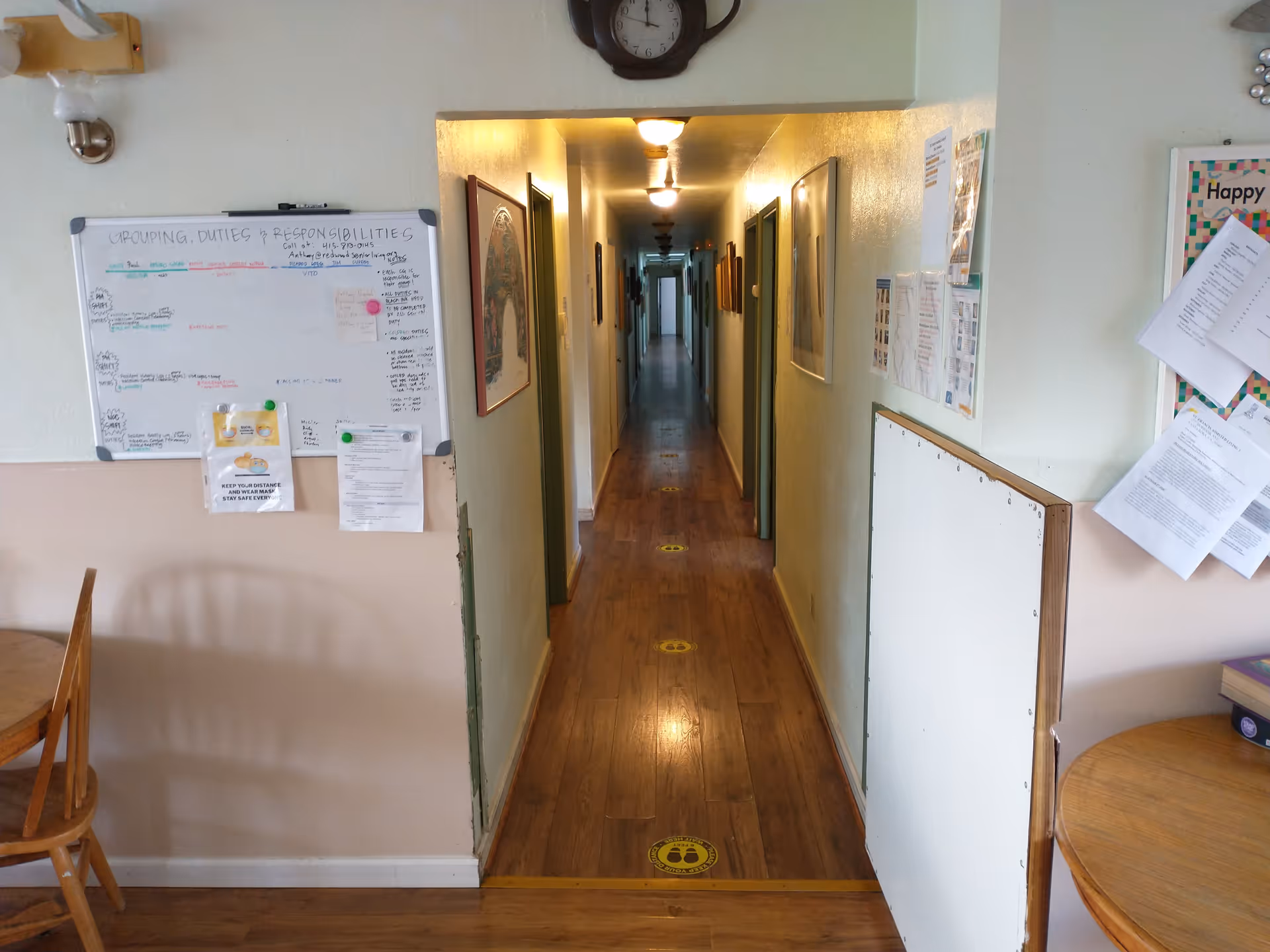 A long, narrow hallway with wooden flooring and several doors on both sides. The hallway is lit by ceiling lights and has framed pictures on the walls. At the entrance to the hallway, there are two round wooden tables with chairs, bulletin boards with papers pinned on the walls, and a whiteboard with notes and reminders. Yellow floor markers are spaced along the hallway floor.