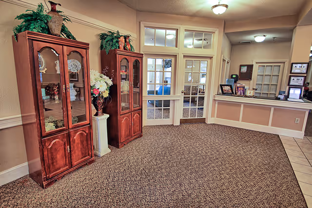 Interior view of a senior living facility lobby area with two wooden display cabinets containing decorative items and plants on top. There is a reception desk on the right side with framed pictures and a computer monitor. The background shows glass-paneled doors leading to another room.